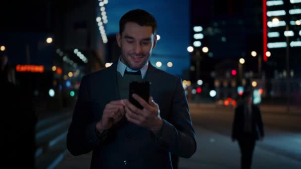Καυκάσιος επιχειρηματίας με κοστούμι χρησιμοποιεί ένα Smartphone στη Σκοτεινή Οδό το βράδυ. Άλλοι άνθρωποι του γραφείου περνούν. Φαίνεται σίγουρος και επιτυχημένος. Ατμοσφαιρική Αστική Πόλη Φώτα στο παρασκήνιο. — Αρχείο Βίντεο