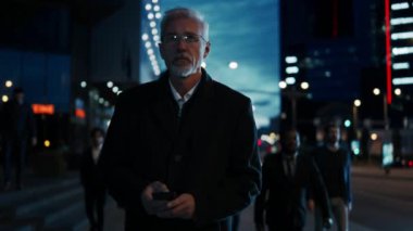 Kıdemli İşadamı Akşamları Dark Street 'te Akıllı Telefon Kullanırken Yürüyor. Ofisteki diğer insanlar yanımızdan geçiyor. Kendinden emin ve başarılı birine benziyor. Atmosferik Şehir Işıkları Arkaplanda.