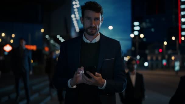 Hombre de negocios caucásico en un traje está utilizando un teléfono inteligente en la calle oscura en la noche. Otras personas de la oficina caminan más allá. Parece confiado y exitoso. Luces urbanas atmosféricas en el fondo . — Vídeo de stock