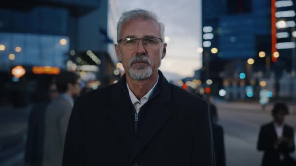 Porträt eines seriösen Geschäftsmannes im Mantel, der auf einer Straße mit Fußgängern steht. Er hat einen Bart und trägt eine Brille. Es ist Abend mit stimmungsvollen urbanen Lichtern im Hintergrund. — Stockvideo