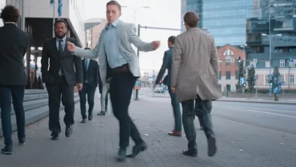 Joven empresario alegre en una chaqueta gris está bailando activamente en una calle de la ciudad. Administrador de la oficina se mueve a través de aglomeraciones de oficinas ocupadas que viajan al trabajo. Centro con rascacielos en el fondo . — Vídeo de stock