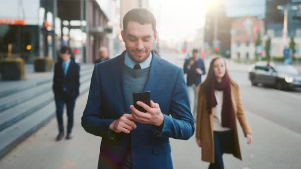 Kaukaski biznesmen w garniturze używa smartfona na ulicy w centrum. Inni ludzie z biura chodzą przeszłością. Uśmiecha się i wygląda na szczęśliwego. Przegląda sieć na swoim urządzeniu.. — Wideo stockowe