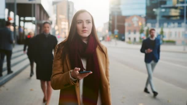 Jeune femme habillée de façon décontractée intelligente utilise un téléphone intelligent dans une rue. Les gens d'affaires et les gestionnaires de bureau passent leur chemin vers le travail. Elle a l'air confiante en vérifiant sa cellule et en marchant . — Video