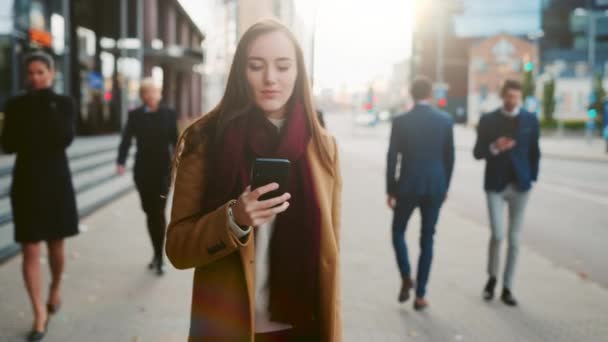 Jovem inteligente Casualmente Vestida Feminino está usando um smartphone em uma rua. Pessoas de negócios e gerentes de escritório caminham em seu caminho para o trabalho. Ela parece confiante ao verificar seu celular e andar . — Vídeo de Stock