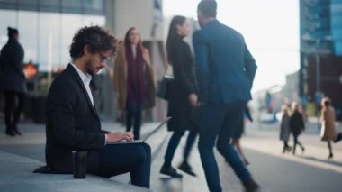 Takım Elbiseli Yakışıklı İş Adamı İş Merkezi 'nin yanındaki basamaklarda oturuyor ve şehirdeki bir sokakta dizüstü bilgisayarda çalışıyor. Ofisteki İnsanlar İşe Yürüyor.