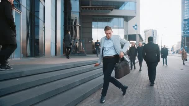 Jovem empresário alegre em um Grey Blazer está ativamente dançando em uma rua da cidade. O gerente do escritório move-se através de multidões ocupadas do escritório que comutam ao trabalho. Ele está segurando uma caixa de couro . — Vídeo de Stock