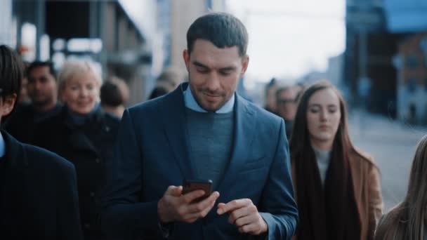 Kaukaski biznesmen w garniturze używa smartfona na ulicy w centrum. Inni pracownicy biurowi dojeżdżają w tłumie. Jest pewny siebie i wygląda na udanego. Przegląda sieć na swoim urządzeniu.. — Wideo stockowe