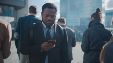 Sokakta yayalarla dikilen takım elbiseli Afrikalı Amerikalı bir iş adamının portresi. O bir Smartphone kullanıyor. Başarılı birine benziyor. Diğer İnsanlar İşe Gidiyor.