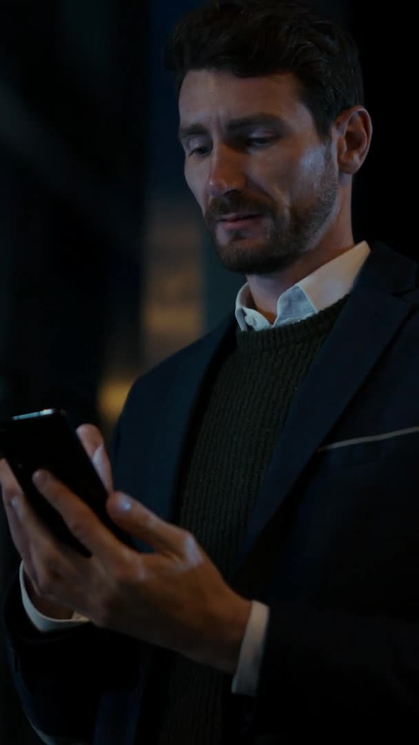 Caucasian Businessman in a Suit is Using a Smartphone on Dark Street in the Evening. Other Office People Walk Past. Video Footage with Vertical Screen Orientation 9:16. — Stock Video