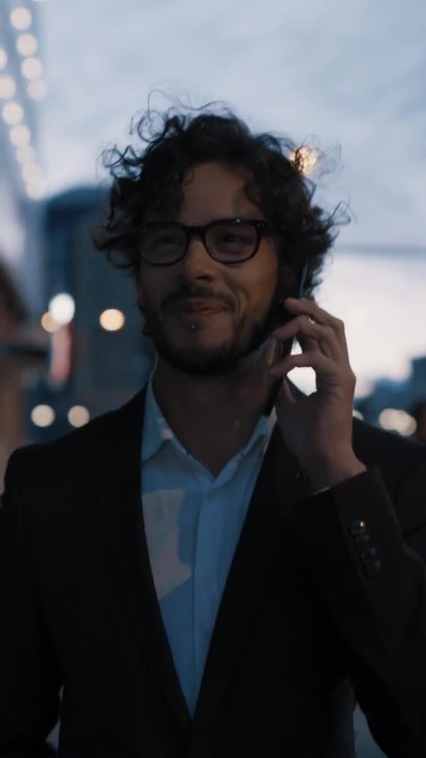 Молодой стильный бизнесмен с кудрявыми волосами разговаривает по телефону во время прогулки по городу. Он в очках. Проходят офисные люди. Видео с вертикальным экраном Ориентация 9: 16 . — стоковое видео