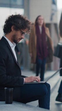 Takım Elbiseli Yakışıklı İş Adamı İş Merkezi 'nin yanındaki basamaklarda oturuyor ve şehirdeki caddede dizüstü bilgisayarda çalışıyor. Ofisteki insanlar işe yürür. Dikey Ekran Yönelimli Video Görüntüsü 9: 16.