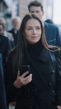 Siyah paltolu güzel iş kadını şehir merkezindeki bir caddede akıllı telefon kullanıyor. Kalabalık bir yaya caddesinde yürüyor ve başarılı görünüyor. Dikey Ekran Yönelimli Video Görüntüsü 9: 16.