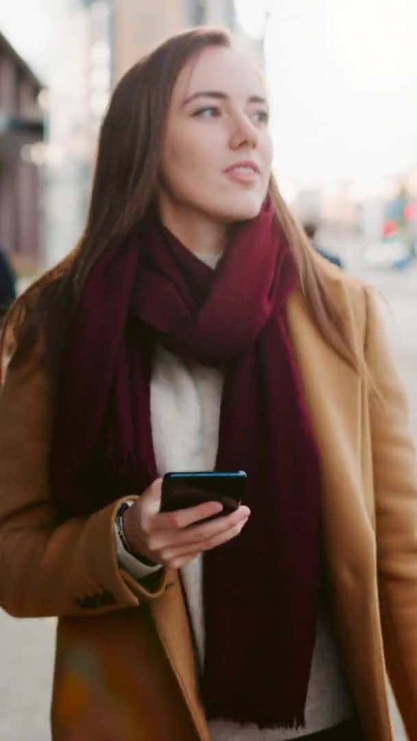 Belle femme d'affaires en manteau brun utilise un smartphone sur une rue du centre-ville. Elle marche sur une rue piétonne bondée et semble réussie. Vidéo avec orientation verticale de l'écran 9 : 16 .