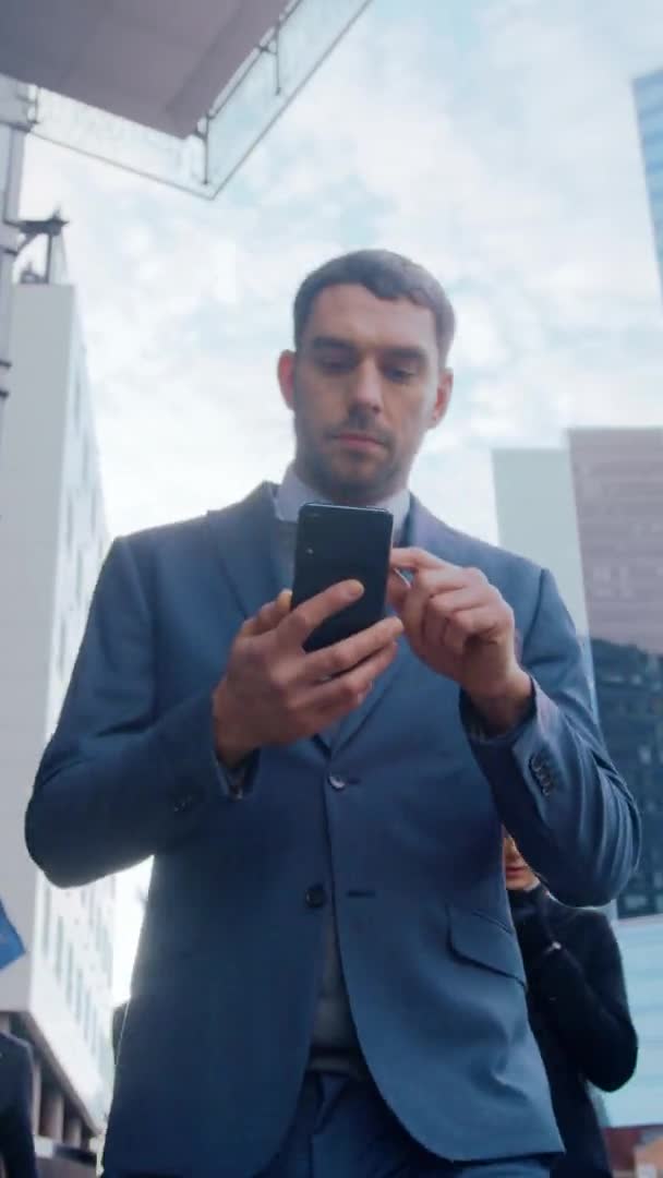 Καυκάσιος επιχειρηματίας με κοστούμι χρησιμοποιεί ένα Smartphone στη Σκοτεινή Οδό το βράδυ. Άλλοι άνθρωποι του γραφείου περνούν. Βίντεο Πλάνα με Κατακόρυφο Προσανατολισμός οθόνης 9: 16. — Αρχείο Βίντεο