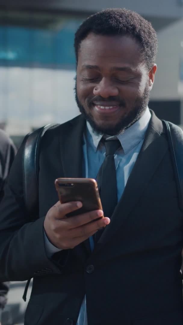Ritratto di un uomo d'affari afroamericano in giacca e cravatta in piedi su una strada con pedoni. Sta usando uno smartphone. Ha un bell'aspetto. Filmati video con orientamento verticale dello schermo 9: 16 . — Video Stock