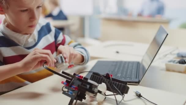 Schoolboy подключает кабели к небольшому роботу, использует ноутбук к программному обеспечению для класса инженерии робототехники. Класс естественных наук начальной школы с одаренными гениальными детьми, работающими с технологиями — стоковое видео
