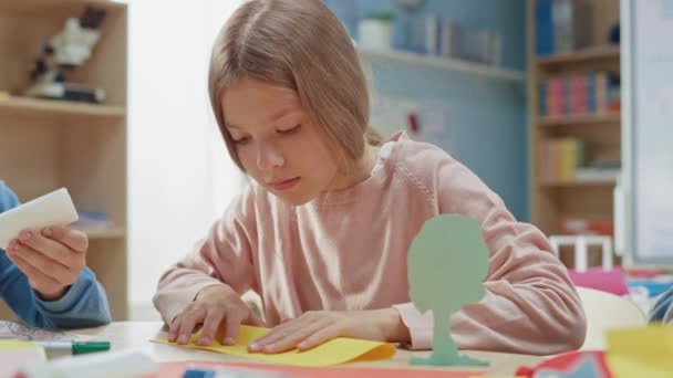 Elementary School Arts & Crafts Class: Linda niña rubia se divierte con sus compañeros de clase en el proyecto de artesanía, utiliza papel colorido para doblar hermoso origami — Vídeos de Stock
