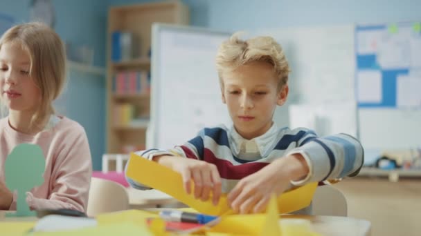 Grundskola konst & hantverk klass: Söt liten lockigt hår pojke använder färgglada papper för att vik vackra Origami, Har kul med sina klasskamrater på hantverk projektet — Stockvideo