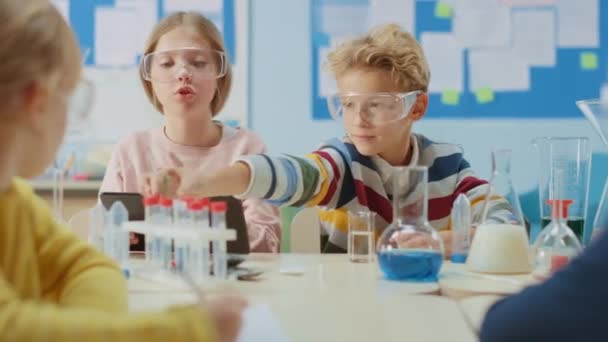 Elementary School Science Classroom: Smart Boy mezcla productos químicos en vasos de precipitados y Cute Girl escribe y analiza los resultados con Tablet Computer digital. Educación moderna, química de aprendizaje — Vídeo de stock