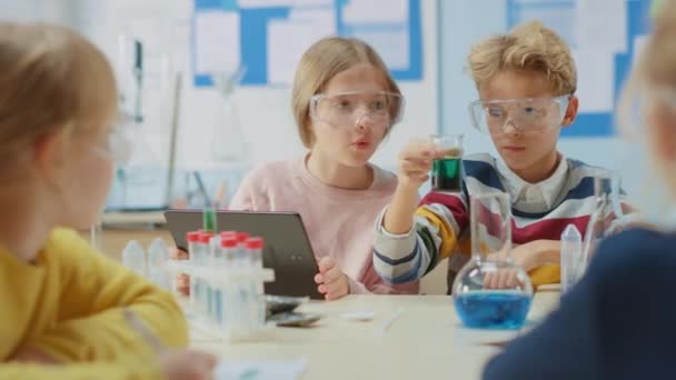 Δημοτικό Σχολείο Science Classroom: Smart Boy αναμιγνύει χημικά σε Μπίκερς και χαριτωμένο κορίτσι γράφει κάτω και αναλύει τα αποτελέσματα με ψηφιακό υπολογιστή tablet. Σύγχρονη Εκπαίδευση, Μαθησιακή Χημεία — Αρχείο Βίντεο