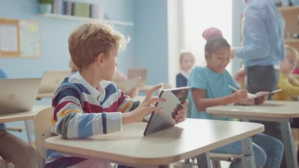 Клас початкової школи комп'ютерних наук: розумний хлопець використовує цифровий планшетний комп'ютер, його однокласники також працюють з ноутбуками. Діти отримують сучасну освіту в STEM, грають і навчаються — стокове відео