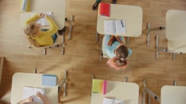 Bright İlköğretim Okulu Sınıfı: Çocuklar okul masasında oturuyor, çalışıyor, ödev yapıyor, Sınırlar Arasında Nazik Öğretmen Ödevleriyle Çocuklara Yardım Ediyor. Dönen Üst Görünüm Kamera Görüntüsü.