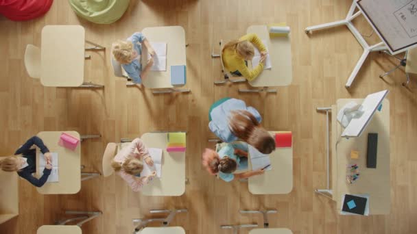 Ljusa grundskolans klassrum: Barn som sitter vid skolskrivbordet och arbetar, gör uppdrag, vänliga lärarpromenader mellan raderna hjälper barn med läxuppgifter. Överst på sidan Kamera Skott. — Stockvideo