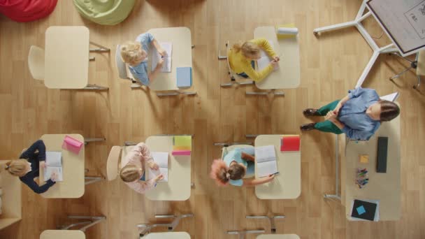 Bright Elementary School Classroom: Children Sitting at their School Desk Working, Doing Assignment, Enthusiastic Teacher Stands a the Head of the Class Explicando uma Lição. Top View Camera Shot . — Vídeo de Stock