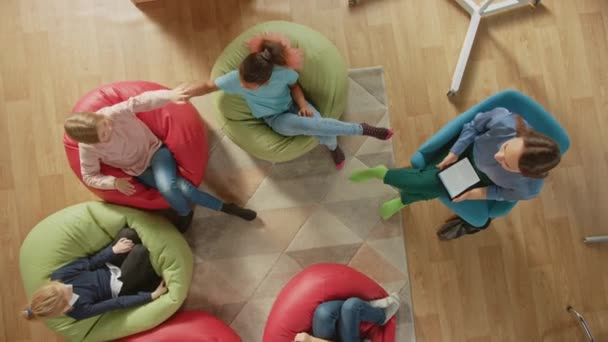 Elementary School Creativity Classroom: Barn sitter på Bean påsar medan läraren förklarar Lesson för dem och använder Digital Tablet Computer, De spelar genom att passera High Five. Ovanifrån kamera — Stockvideo