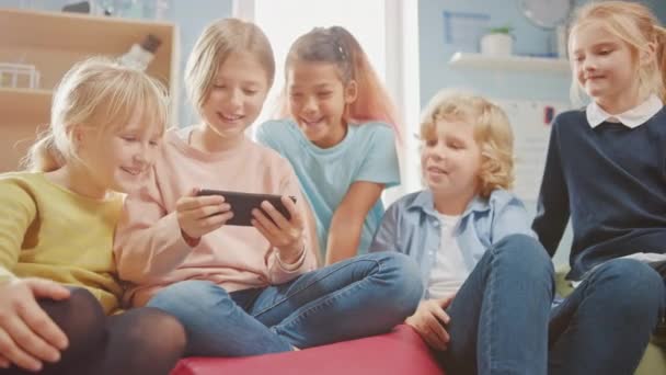 Bambini che navigano su Internet e giocano ai videogiochi online sul telefono cellulare, guardando video. Diversi gruppi di bambini piccoli e carini seduti insieme sulle borse a fagiolo usano smartphone e parlano, divertiti — Video Stock