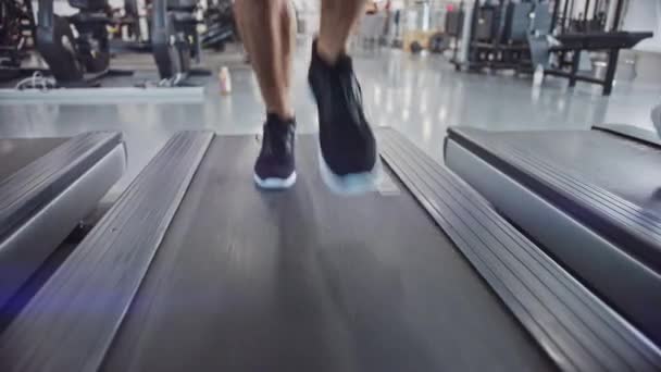 Atletický svalnatý muž běžící na běžeckém pásu, noze a kardio den. Silný mužský trénink v moderním tělocvičně. Cvičení sportovců ve Fitness Clubu. Pohled zepředu na nízkopozemní záběr v pomalém pohybu. Zaměření na nohy — Stock video