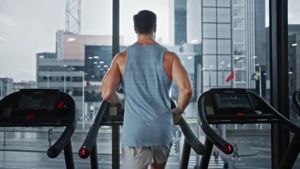 Athletic Muscular Man Correndo em uma esteira, perna e Cardio Day. Forte Homem Treinamento no Ginásio Clube de Fitness Moderno com Janela Vista Cityscape. Câmera de movimento lento vista traseira tiro — Vídeo de Stock