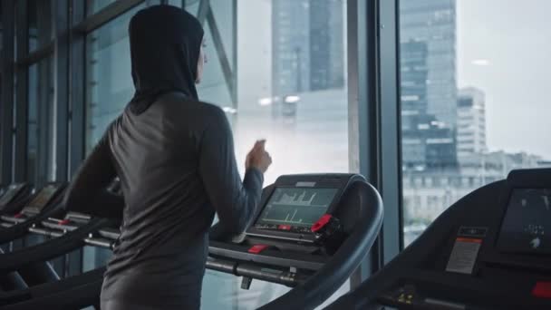 Sportliche muslimische Sportlerin in Hijab und Sportbekleidung, die auf einem Laufband läuft. Energetic Fit Athletin Training allein in der Turnhalle. Blick aus dem Fenster des städtischen Geschäftsviertels. Porträt von der Seite — Stockvideo