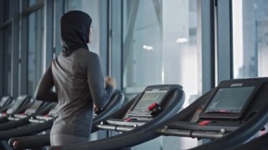 Koşu bandında koşan tesettürlü ve spor giyinen Müslüman Atletik Spor Kadını. Sadece Spor Salonu 'nda Enerjik Formda Kadın Sporcu Eğitimi. Kentsel İş Bölgesi Pencere Manzarası. Yan Görünüm Yavaş Hareket