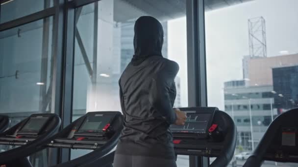 Atletische moslim sportvrouw draagt hidjab en sportkleding rennend op een loopband. Energetische fit vrouwelijke atleet training in de sportschool alleen. Urban Business District Window View. Terug Bekijk Zoom in — Stockvideo