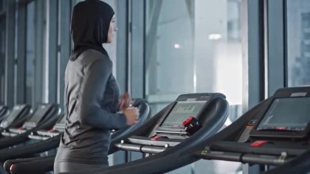 Donna sportiva musulmana atletica che indossa Hijab e abbigliamento sportivo Corsa su un tapis roulant. Allenamento di atleta donna in forma energetica nella palestra da sola. Vista sulla finestra del distretto degli affari urbani. Ritratto vista laterale — Video Stock