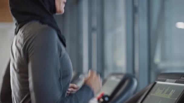 トレッドミルでヒジャブとスポーツウェアを身に着けているアスレチックイスラム教徒のスポーツ女性。ジム単独でのエネルギーフィット女性アスリートトレーニング。都市ビジネス地区ウィンドウビュー。サイドビューポートレートスローモーション — ストック動画