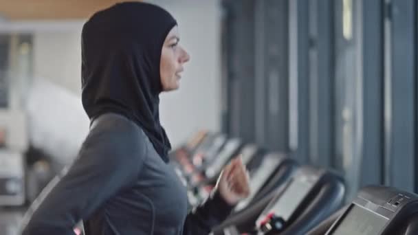 运动的穆斯林女运动员穿着Hijab和运动服在跑步机上跑步。单独在健身房进行精力充沛的女运动员训练。市区商业区窗口视图.侧视图肖像慢动作 — 图库视频影像