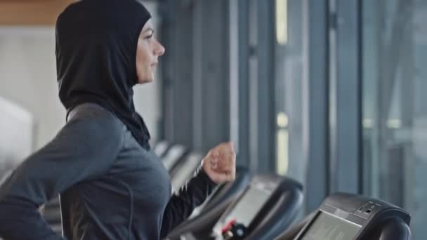 トレッドミルでヒジャブとスポーツウェアを身に着けているアスレチックイスラム教徒のスポーツ女性。ジム単独でのエネルギーフィット女性アスリートトレーニング。都市ビジネス地区ウィンドウビュー。バックビュー｜アークショット — ストック動画