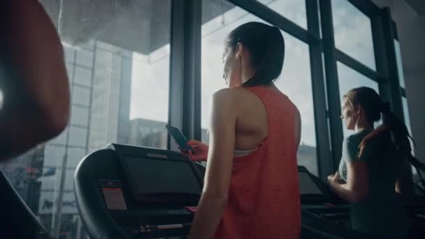 Bella donna sportiva atletica in palestra Correre su un tapis roulant, utilizza lo smartphone. In background si adattano alla formazione degli atleti. Colpo ad arco a basso angolo con movimento lento. Colori freddi scuri — Video Stock