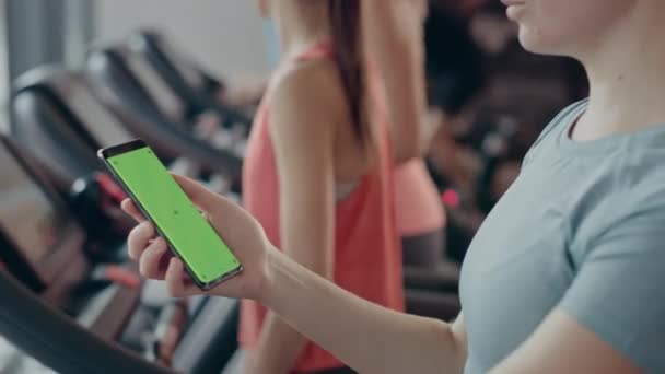 Mujer atlética utiliza Green Mock-up Screen Smartphone mientras se ejecuta en la cinta de correr en un gimnasio. Deportes Mensajes femeninos en las redes sociales, toma fotos, ve videos mientras hace ejercicio en el club de fitness — Vídeos de Stock