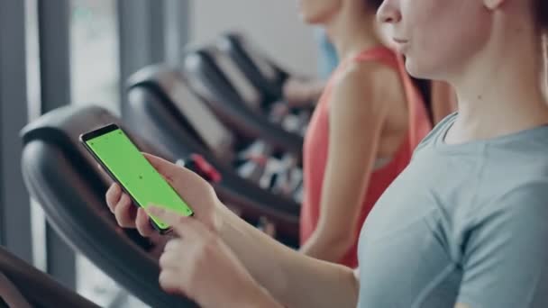 Sportowa kobieta robi gesty podczas korzystania z zielonego ekranu smartfona i bieganie na bieżni w siłowni. Fit Kobieta Posty w mediach społecznościowych, Robi zdjęcia, Ogląda filmy, Ćwiczenia w klubie fitness — Wideo stockowe