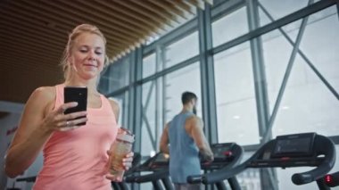 Güzel Atletik Kadın Spor Salonunda Akıllı Telefon Kullanıyor. Sosyal medyayı kullanmak, resimleri yüklemek, iletişim kurmak, e-postaları kontrol etmek. Arka planda Koşu bandında Koşan İnsanlar