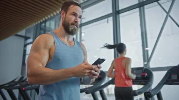 Kas Ağırsiklet Şampiyonu Spor Salonunda Yürüyor, Sosyal Medya ve İş İlişkileri için Akıllı Telefon kullanıyor. Arka planda Koşu bandında Koşan İnsanlar — Stok video