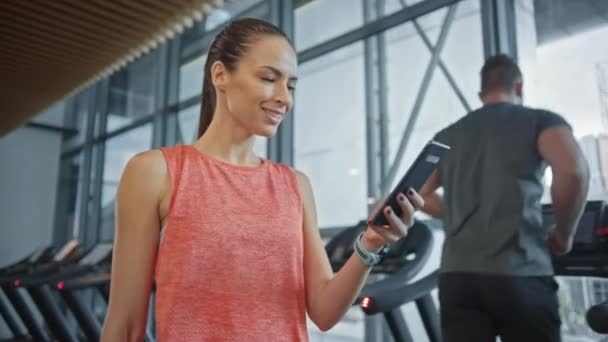 Прекрасна спортсменка використовує смартфон "Walks Through Fitness Club Gym". Використовуючи соціальні медіа, Posting Pictures, Communicating, Checking Email. На задньому плані спортивні люди бігають по Тредміллерах — стокове відео