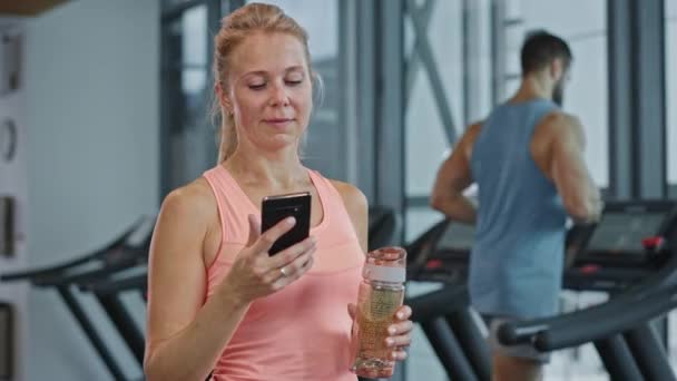 Krásná atletická žena používá Smartphone procházky Fitness Club tělocvična. Používání sociálních médií, účtování obrázků, komunikace, kontrola e-mailu. V pozadí Sport Lidé běží na běžeckých pásech — Stock video