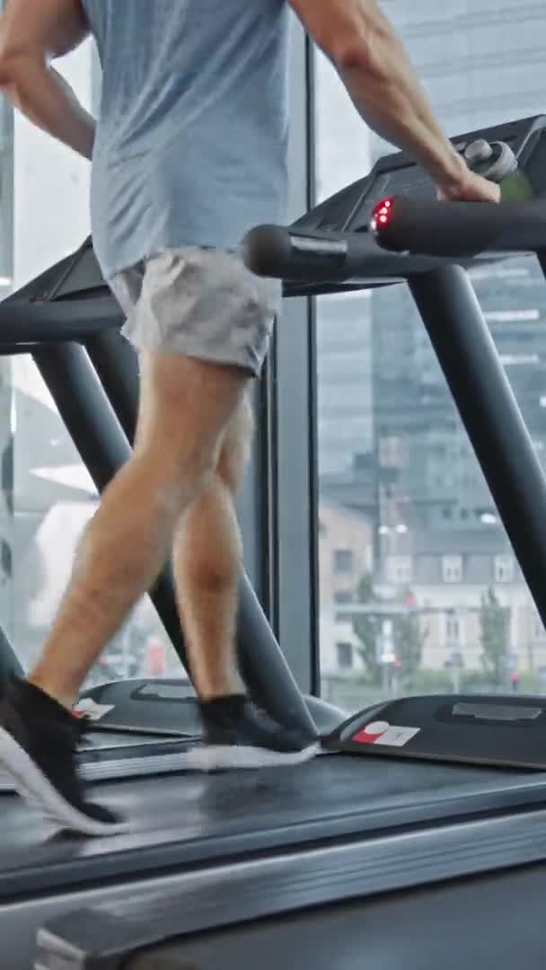 Atletický svalnatý muž běžící na běžeckém pásu, noze a kardio den. Silný mužský trénink v Moderní tělocvičně. Pohled zepředu na nízkopozemní záběr. Soustřeď se na nohy. Svislá orientace obrazovky Video 9: 16 — Stock video