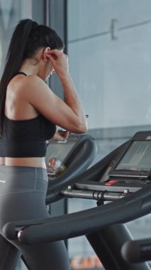 Spor salonundaki Güzel Spor Kadını Kablosuz Kulaklıkla Koyuyor, Podcast 'i açıyor ya da Smartphone ile Spor Müziği Çalıyor ve Koşu bandında Başlıyor. Dikey Ekran Yönlendirme Videosu 9: 16