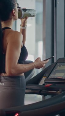 Koşu bandında Koşan Güzel Spor Kadını Akıllı Telefon ve Tumbler 'dan Protein Ek Sıvısı Sıvısı Kullanıyor. Dikey Ekran Yönlendirme Videosu 9: 16