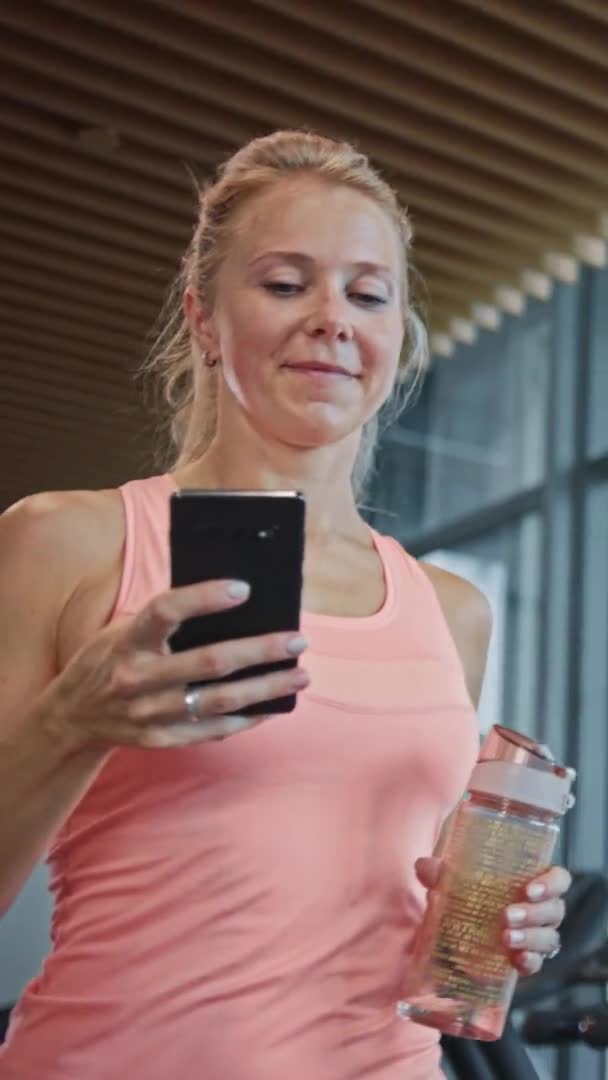 Mulher Atlética bonita usa Smartphone Walks Through Fitness Club Gym. Usando mídias sociais, postando imagens, se comunicando, verificando e-mail. Vídeo de Orientação de Tela Vertical 9: 16 — Vídeo de Stock